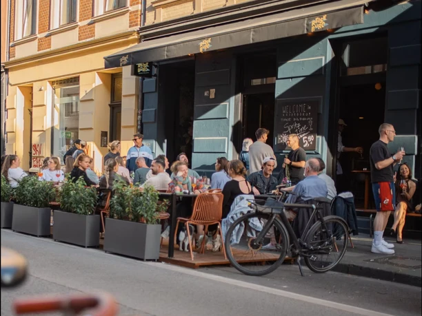 Reisebüro Terrasse Menschen sitzen und stehen vor einer Bar mit Außenterrasse in einer belebten Straßenszene