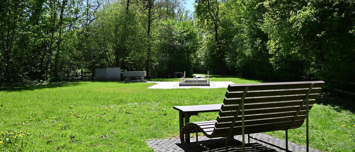 Holzbank vor Kneipptretbecken auf sonniger Wiese, umgeben von hohen Bäumen.