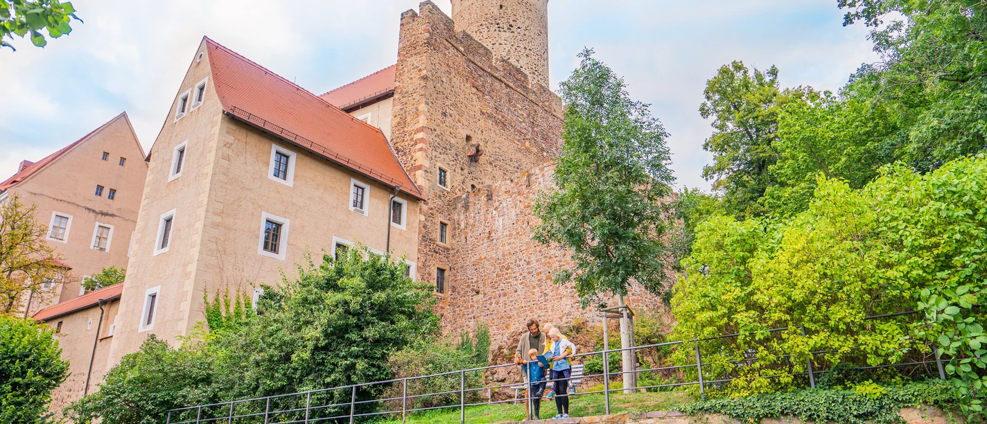 Zu sehen ist die Burg Gnandstein in einer Nahaufnahme. Im Vorgrund ist eine Familie mit zwei Kinder zu erkennen, die sich umschaut.