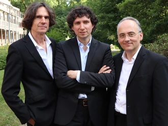 Das Trio "Ocean´s 3" Drei Musiker in schwarzen Anzügen posieren im Grünen vor einem Gebäude.