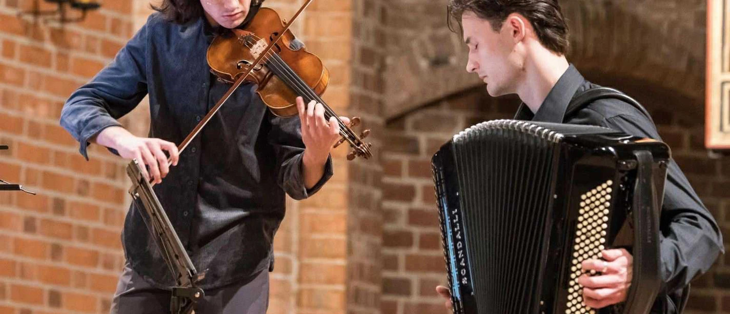 Kammermusikgruppe Profunduo Zwei Musiker von Profunduo spielen Violine und Akkordeon vor Backsteinmauer.