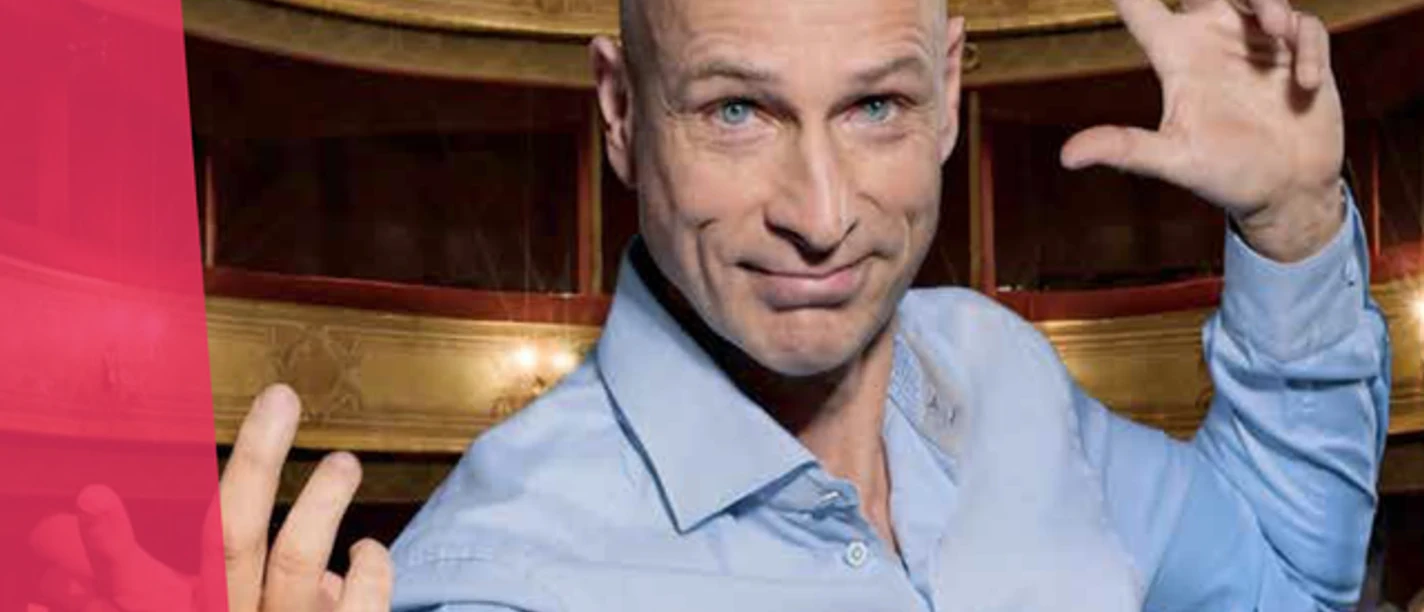 JC Scheibe Glatzköpfiger Mann im Theater hebt die Hände, im Hintergrund Zuschauer und Noten.