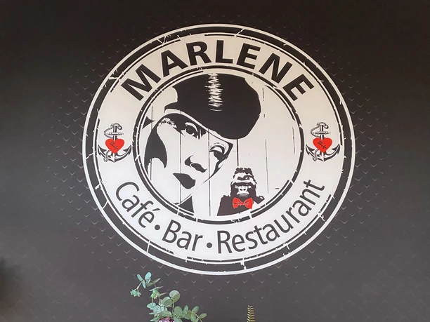 IMG_0773 (c) Nicole Schillig_Erlebnis Bremerhaven_WEB.jpg Rundes Logo des Marlene Café Bar Restaurants über einer schwarzen Kommode mit Flaschen und Blumen.