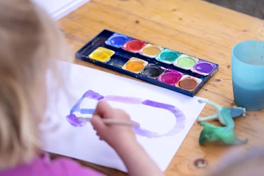 Mit Wasser malen Kind malt mit Wasserfarben auf Papier, daneben Farbkasten, Becher und kleines Spieltier.