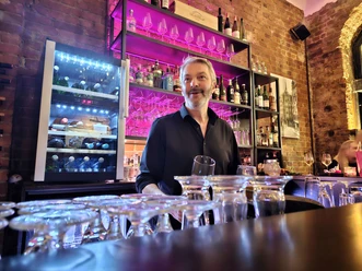 Freundlicher Barkeeper am Tresen vor Backsteinwand mit pink beleuchtetem Regal.