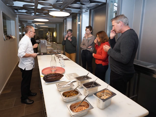 Cafe Amrein Chocolatier_Choco Workshop Teilnehmende eines Choco Seminars im Cafe Amrein Willisau stehen an einer langen Arbeitsfläche und probieren verschiedene Schokoladen und Kakaoerzeugnisse während
