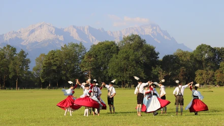 Trachtenverein Oberau Trachtengruppe tanzt auf einer Wiese vor Baumreihe und hellen Berggipfeln im Sonnenlicht