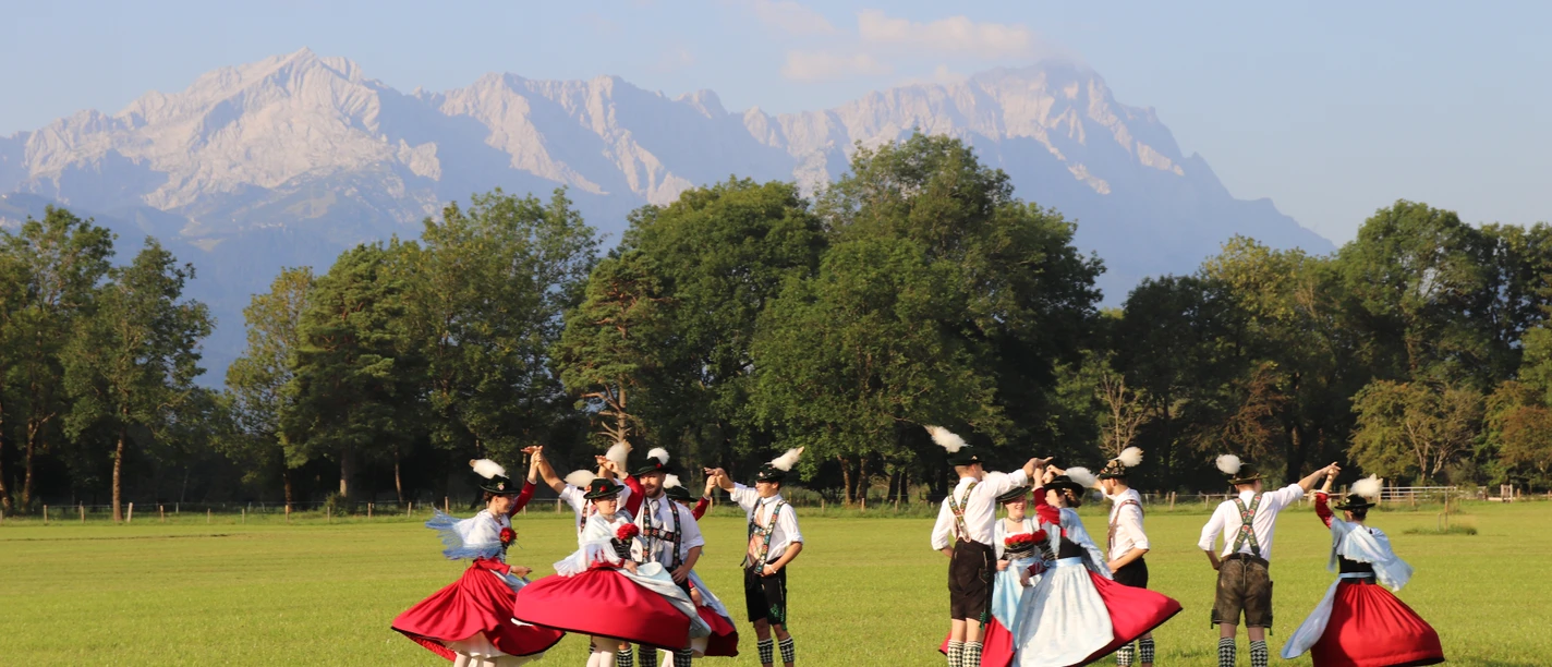 Trachtenverein Oberau Trachtengruppe tanzt auf einer Wiese vor Baumreihe und hellen Berggipfeln im Sonnenlicht