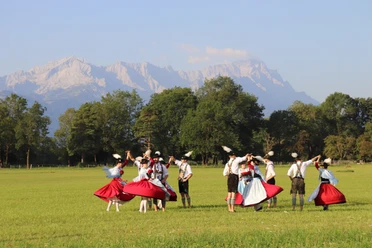 Trachtenverein Oberau Trachtengruppe tanzt auf einer Wiese vor Baumreihe und hellen Berggipfeln im Sonnenlicht