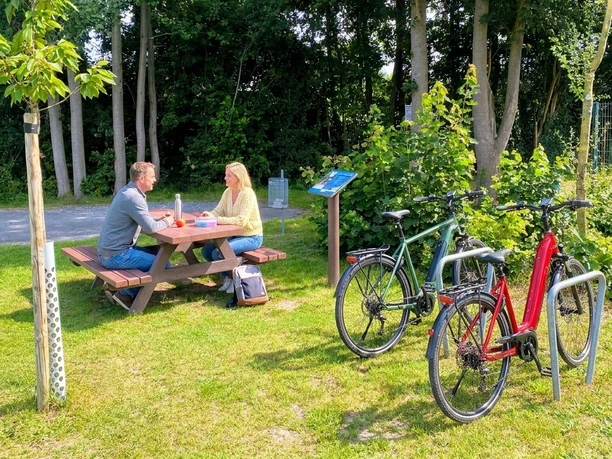Rastplatz mit Picknicktisch, zwei Radfahrende pausieren neben abgestellten Fahrrädern im Grünen
