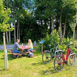 Rastplatz Ströhen (C) Gemeinde Steinhagen - KI-gestützte Bildbearbeitung.jpg Rastplatz mit Picknicktisch, zwei Radfahrende pausieren neben abgestellten Fahrrädern im Grünen