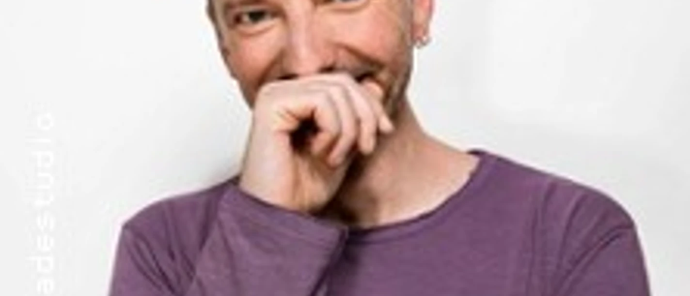 Mann mit kurzem Haar in violettem Shirt lächelt und hält eine Hand vor den Mund.