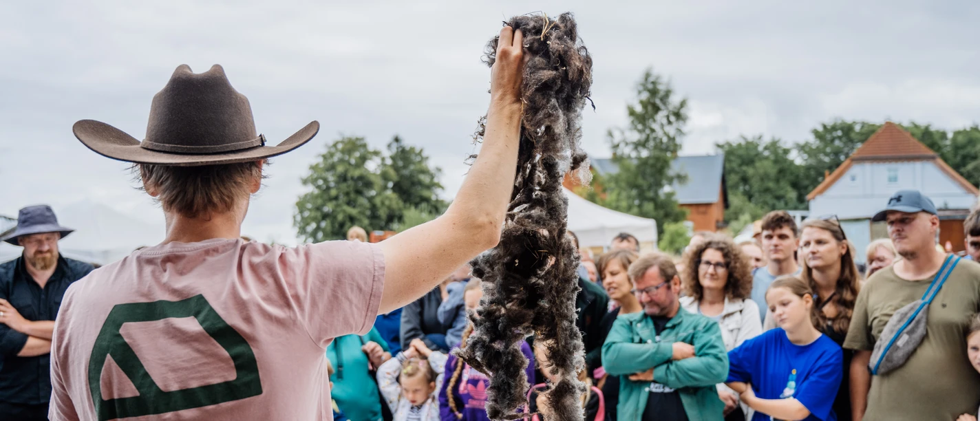 Nordwolle Werksfestival Person mit Hut präsentiert eine dunkle Wollfaser vor einer dicht versammelten Zuschauergruppe.