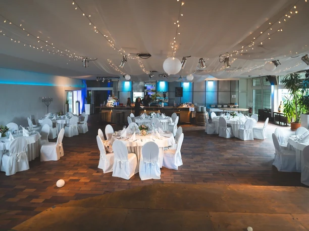Tafelfreuden A festively decorated hall for a banquet, featuring round tables, string lights, and a bar