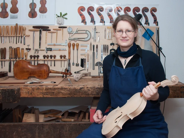 MeineLGeigen - Master violin maker Anna Karoline Meinel - Regional products from Leipzig and the region Master violin maker Anna Karoline Meinel sits in her workshop and holds a white finished violin in front of her tool wall.