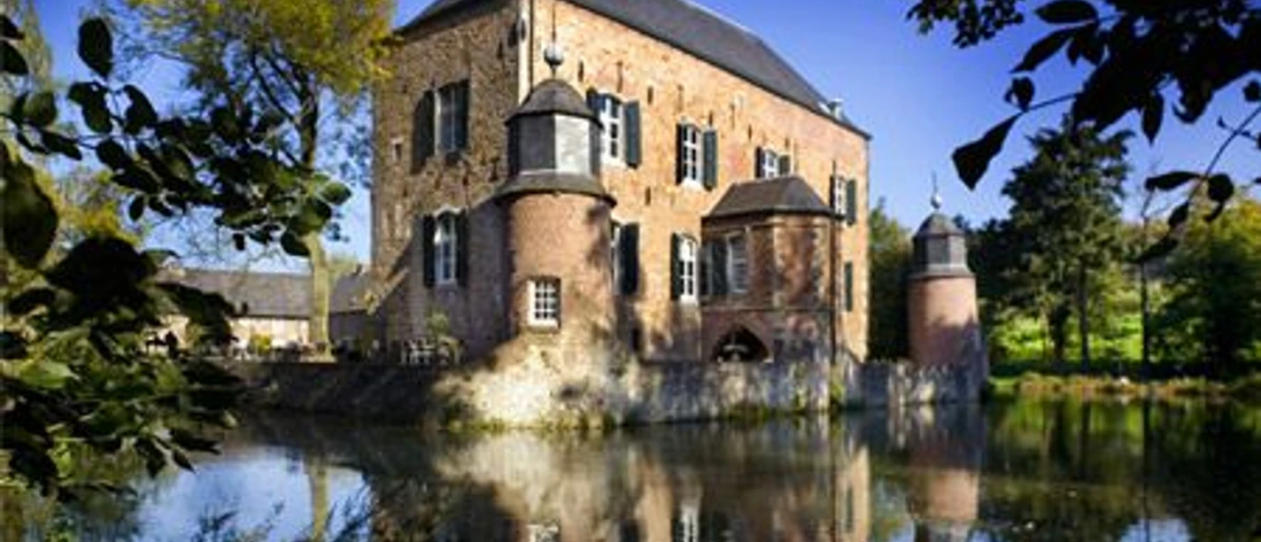 Außenansicht Kasteel Erenstein Schloss mit zwei Türmern rechts und links, die niedriger sind als das restliche Gebäude. Es liegt in einem See und ringsherum sind Bäume.
