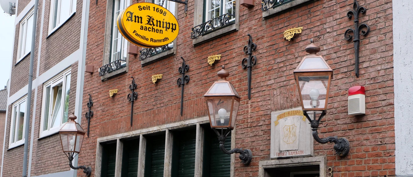 Am Knipp Aachen Außenansicht und Fassade des Restaurants "Am Knipp". Sie ist geschmückt mit alten Laternen und Verzierungen. Zwischen den Fenstern des Erdgeschosses und der ersten Etage hängt ein Schild an einer Halterung, auf dem der Name des Restaurants steht.