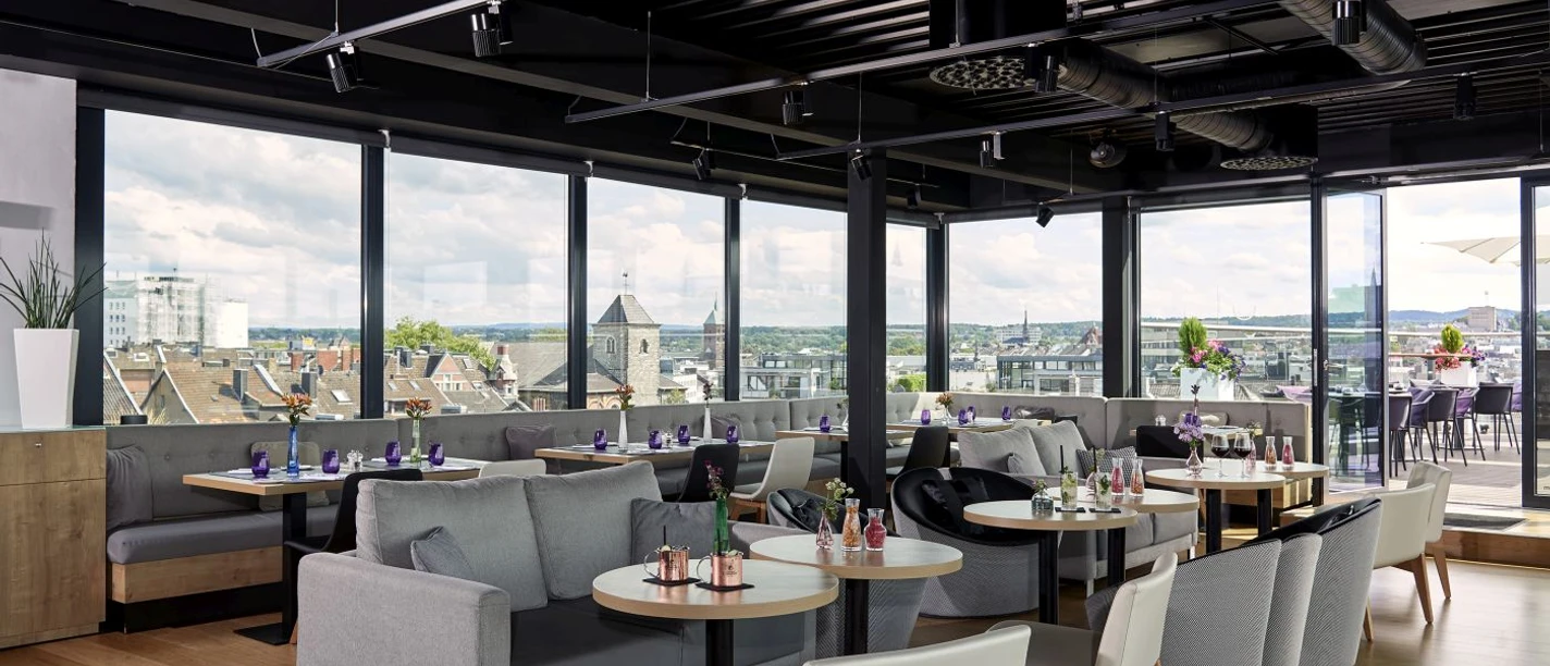 Innside Aachen Up Town Bar Lounge Raum mit Sofas, runden Tischen und Stühlen und Fensterfront mit Blick über Aachener Dächer.