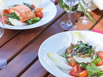 Restaurant Mediterraneo KOCHKULTUR Nahaufnahme von einem Holztisch mit leeren Weingläsern und zwei Tellern. Auf dem einen Teller ist gekochter Lachs mit Salat und auf dem anderen ein Salat mit Ziegenkäse und Chicoree.