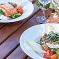 Restaurant Mediterraneo KOCHKULTUR Nahaufnahme von einem Holztisch mit leeren Weingläsern und zwei Tellern. Auf dem einen Teller ist gekochter Lachs mit Salat und auf dem anderen ein Salat mit Ziegenkäse und Chicoree.