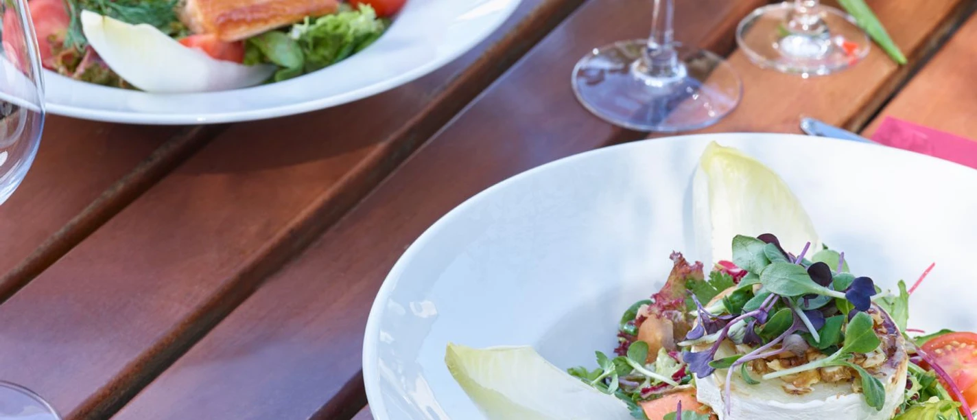 Restaurant Mediterraneo KOCHKULTUR Nahaufnahme von einem Holztisch mit leeren Weingläsern und zwei Tellern. Auf dem einen Teller ist gekochter Lachs mit Salat und auf dem anderen ein Salat mit Ziegenkäse und Chicoree.