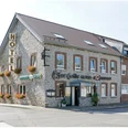 Außenansicht des Restaurants "Zur Heide" mit einer Fassade aus Bruchstein. Das Eckhaus grenzt an eine Straßenkreuzung und ist zweistöckig. An den Fenstern auf der kurzen Seite befinden sich Blumenkübel und vertikal angebrachte Schilder mit den Buchstaben "HOTEL". An der langen Seite des Restaurant befinden sich ebenfalls Fenster, die Aufschrift "Zur Heide Hotel Restaurant" sowie der überdachte Eingang. und