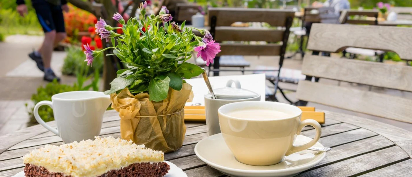 Ein runder Holztisch und ein Holzstuhl stehen draußen. Auf dem Tisch steht eine Kaffeetasse auf einer Untertasse, eine kleine Kanne mit Milch und eine mit Zucker, ein Blumentopf und ein Teller mit einem Stück Kuchen. Im Hintergrund lassen sich weitere Tische und Stühle erahnen. Drumherum ist es begrünt.