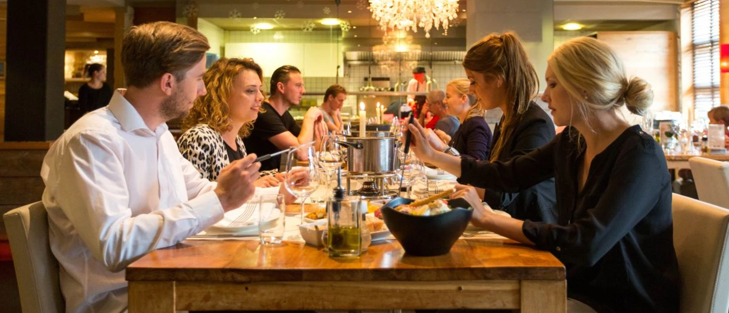 Mehrere Menschen sitzen an einem gedeckten Holztisch mit Stabkerzen und Töpfen in der Mitte und essen Käse-Fondue. Von der Decke hängen zwei Kronleuchter.