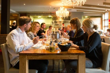 Fonduerestaurant - Snowworld Landgraaf Mehrere Menschen sitzen an einem gedeckten Holztisch mit Stabkerzen und Töpfen in der Mitte und essen Käse-Fondue. Von der Decke hängen zwei Kronleuchter.