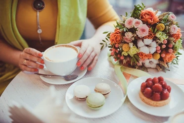 Runder Café-Tisch mit einem bunten Blumenstrauß, einem Erdbeertörtchen, drei Macarons und einem Cappuccino, den eine Frau mit der Hand festhält.