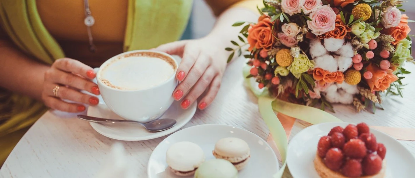 Aachener Café Haus Runder Café-Tisch mit einem bunten Blumenstrauß, einem Erdbeertörtchen, drei Macarons und einem Cappuccino, den eine Frau mit der Hand festhält.
