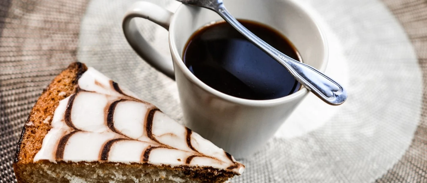 Nahaufnahme von einem schwarzen Kaffee in einer Tasse, auf der ein Löffel abgelegt ist und einem Stück Kuchen.