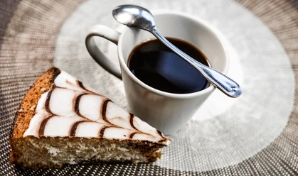 Café M Nahaufnahme von einem schwarzen Kaffee in einer Tasse, auf der ein Löffel abgelegt ist und einem Stück Kuchen.