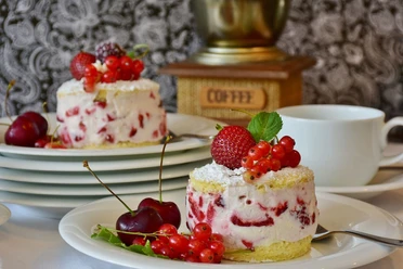 Nahaufnahme von einem Teller, auf dem ein rundes Stück Kuchen mit Erdbeeren und Johannisbeeren steht. Im Hintergrund stehen gestapelte Teller, wobei auf dem oberen auch ein solches Kuchenstück liegt.