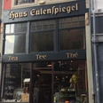 Der Teeladen - Haus Eulenspiegel Dunkle Fassade mit der Aufschrift "Haus Eulenspiegel" und "Tea", "Tee" und "Thé" darunter. Hinter dem Schaufenster im Erdgeschoss ist Ware ausgestellt. Der Eingang befindet sich in der Mitte.