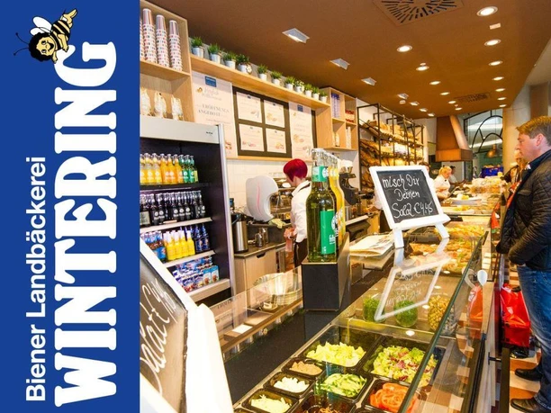 Wintering Innenansicht einer modernen Bäckerei mit Verkaufstheke, frischen Backwaren und Kundschaft beim Einkauf.