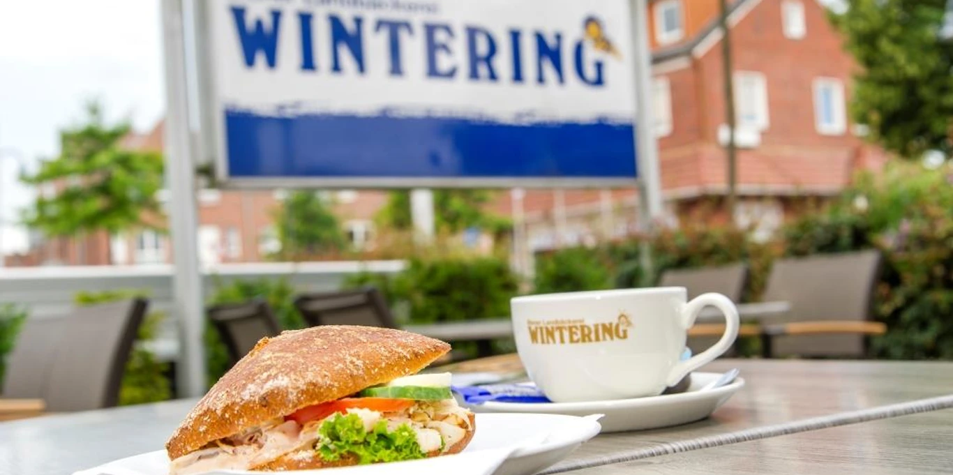 Wintering_Spelle_klein.jpg Ein belegtes Brötchen und eine Kaffeetasse mit Wintering-Logo auf einem Tisch vor dem Café in Spelle