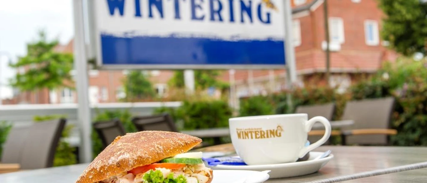 Wintering_Spelle_klein.jpg Ein belegtes Brötchen und eine Kaffeetasse mit Wintering-Logo auf einem Tisch vor dem Café in Spelle