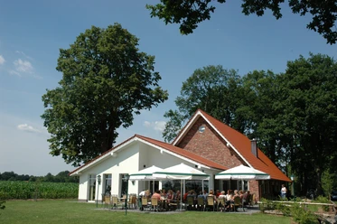 Backsteinhaus mit rotem Dach, Sonnenschirmen und Gästen auf der Terrasse in grüner Umgebung.