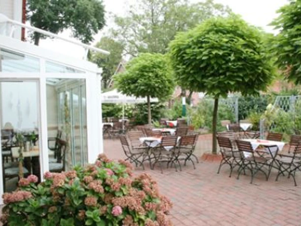 Gasthof_Hense_-_Cafe.JPG Außenbereich eines Cafés mit Holztischen und Stühlen unter schattigen Bäumen auf gepflasterter Terrasse.