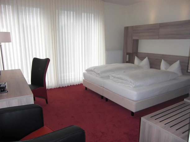 Hotel_Altes_Landhaus_Lingen_-_Zimmer_mini_01.jpg Helles Hotelzimmer mit Doppelbett, rotem Teppich, Holzmöbeln, Schreibtisch und bodentiefem Fenster