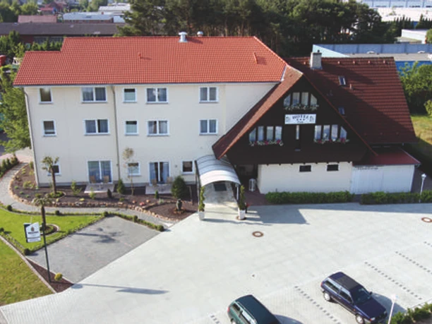 Hotelgebäude mit rotem Satteldach, hellem Anbau und überdachtem Eingang vor breiter Parkfläche