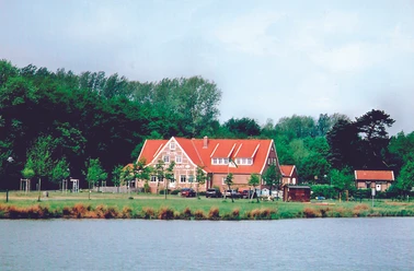 Backsteinbau mit rotem Satteldach am Waldrand, davor Wiese, parkende Autos und ein See im Vordergrund.