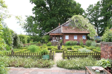 Backsteinhaus mit Satteldach, umgeben von grünem Kräutergarten und Holzzaun unter alten Bäumen.