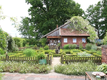 kr-uterhof-rosen5-emsland-touristik-birgit-janknecht Backsteinhaus mit Satteldach, umgeben von grünem Kräutergarten und Holzzaun unter alten Bäumen.