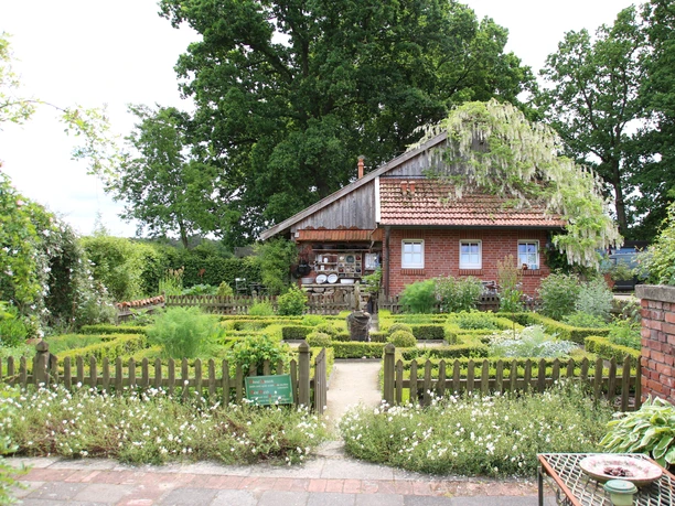kr-uterhof-rosen5-emsland-touristik-birgit-janknecht Backsteinhaus mit Satteldach, umgeben von grünem Kräutergarten und Holzzaun unter alten Bäumen.