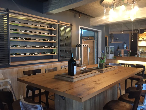 deichkrone-johanna-l-bbers-13 Holztisch mit Stühlen, Weinflaschenregal an der Wand und warm beleuchtetem Tresen im Hintergrund