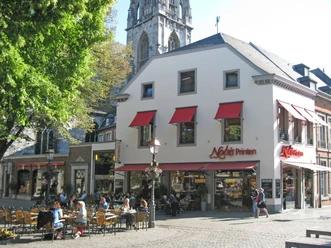 Nobis Printen Münsterplatz Auf dem Münsterplatz befindet sich das große Haus der Bäckerei Nobis mit vielen Sitzgelegenheiten davor, die zur Hälfte mit Kunden belegt sind. Links ragt ein blühender Baum ins Bild.