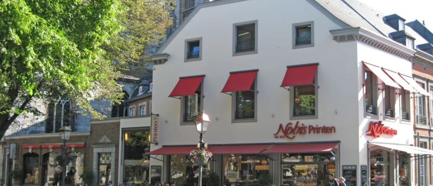 Nobis Printen Münsterplatz Auf dem Münsterplatz befindet sich das große Haus der Bäckerei Nobis mit vielen Sitzgelegenheiten davor, die zur Hälfte mit Kunden belegt sind. Links ragt ein blühender Baum ins Bild.
