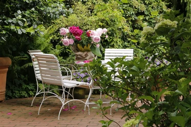 regionales-emsland-touristik-birgit-janknecht-3 Weiß lackierte Gartenstühle und ein Tisch mit Blumenstrauß aus Pfingstrosen zwischen dichtem Grün.